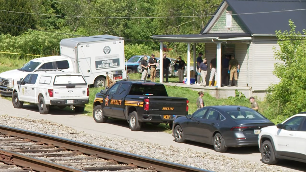 Sheriff's vehicles and a Crime Scene Unit trailer parked near a house where law enforcement personnel are gathered on the porch.