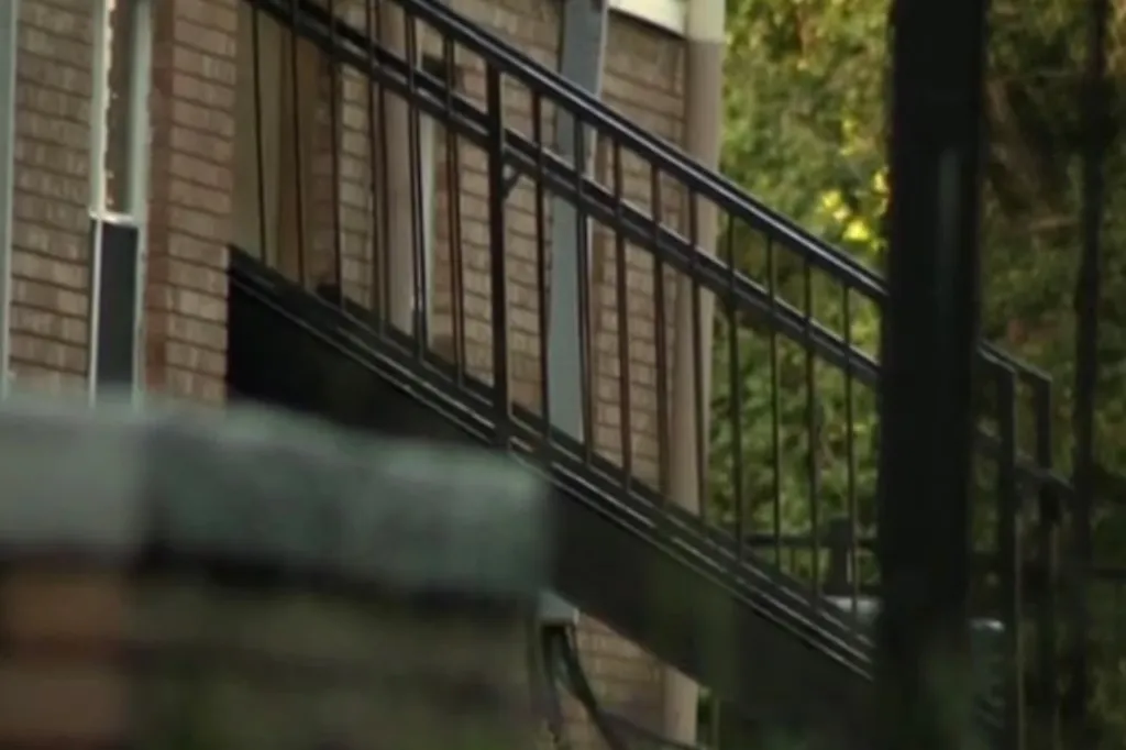 Exterior view of a building with a brown brick facade and dark metal railing of an outdoor staircase or balcony.