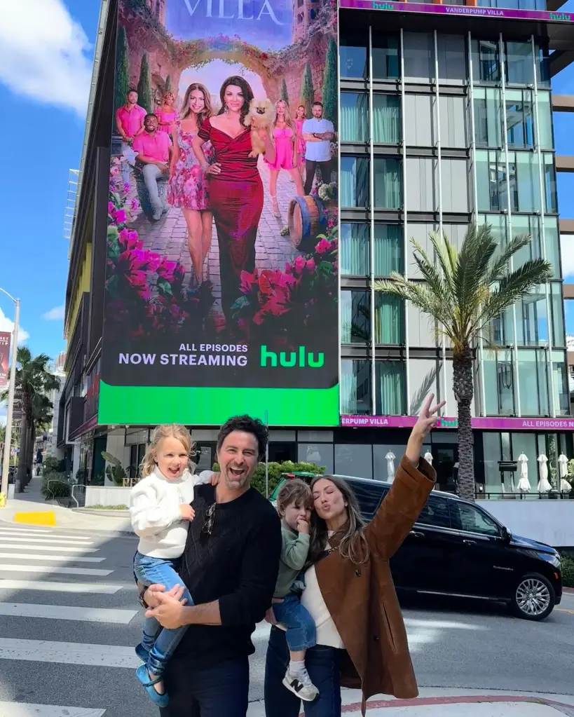 Stassi Schroeder, her husband Beau, and their two young children pose in front of a large "Vanderpump Villa" Hulu billboard in a city.