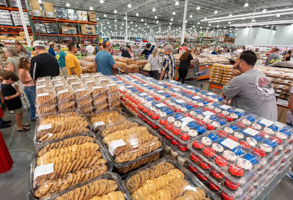 People shop in the bakery section of a Costco, with large displays of cookies and red, white, and blue cupcakes in the foreground.
