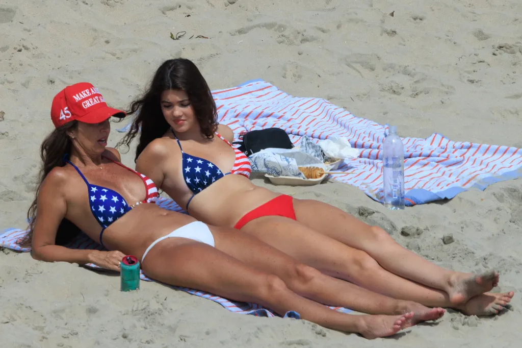 Two women in American flag bikinis relax on a beach towel on the sand.