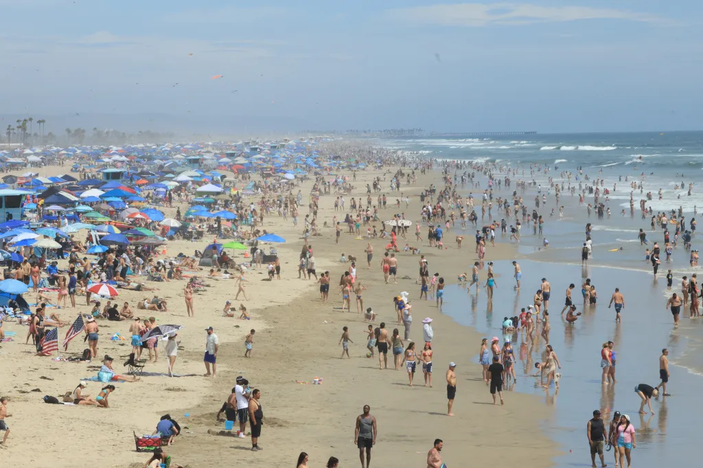 Crowds of people gather on Huntington Beach in California, with many under colorful umbrellas on the sand and others in the shallow water.