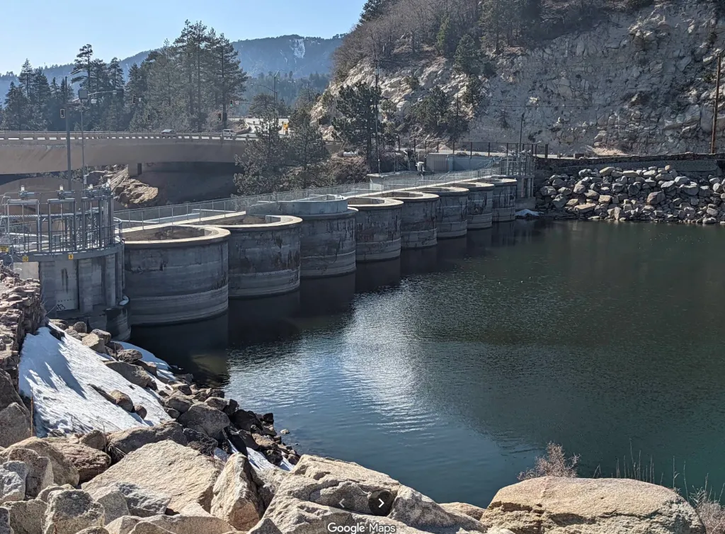 The Bear Valley Dam with a concrete bridge overhead and a mountainous, treed landscape in the background.