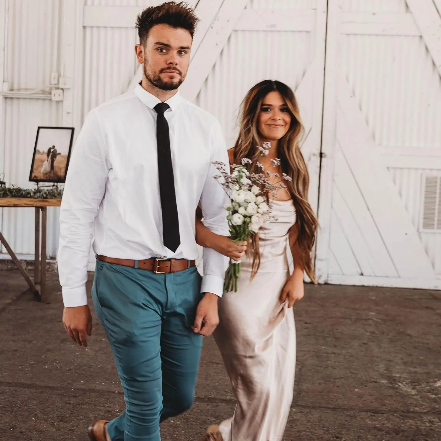 A man in a white shirt and tie walking arm-in-arm with a woman in a satin dress holding a bouquet.