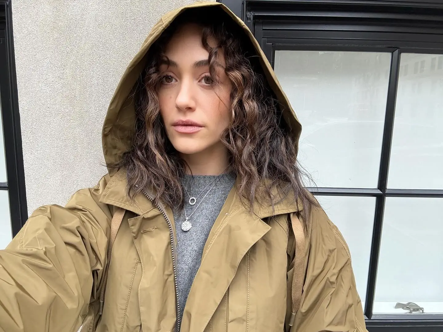 Emmy Rossum wearing a hooded tan jacket with a gray sweater.