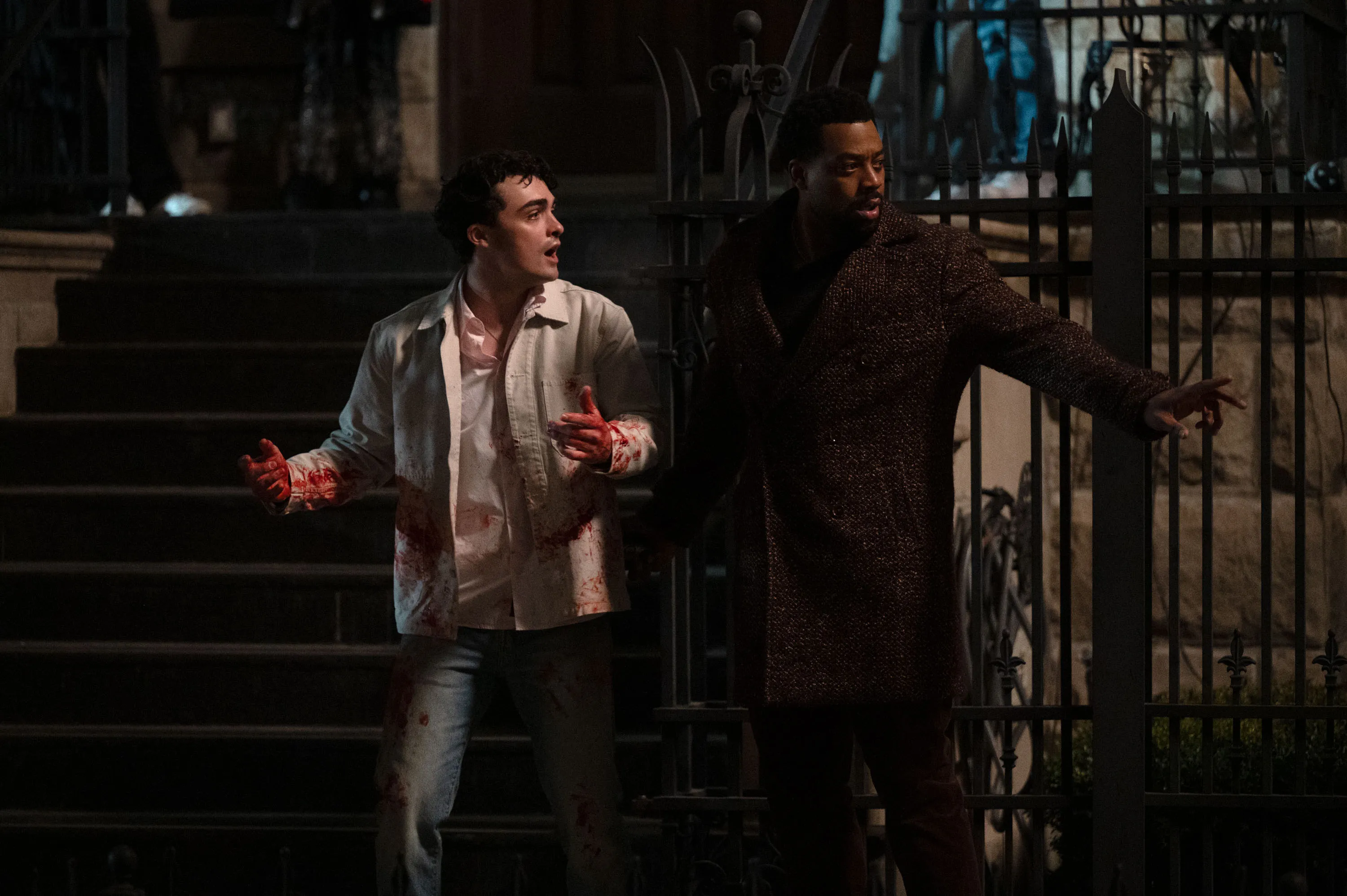 Ben Larson as Ellis, with blood on his shirt and hands, and LaRoyce Hawkins as Officer Kevin Atwater, standing next to him, with dark stairs behind them and an iron fence.
