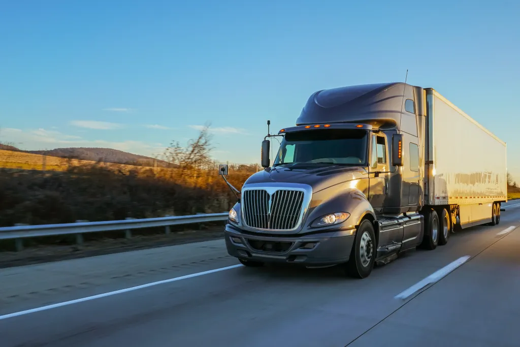 A gray semi-truck with a white trailer drives on a highway at golden hour.