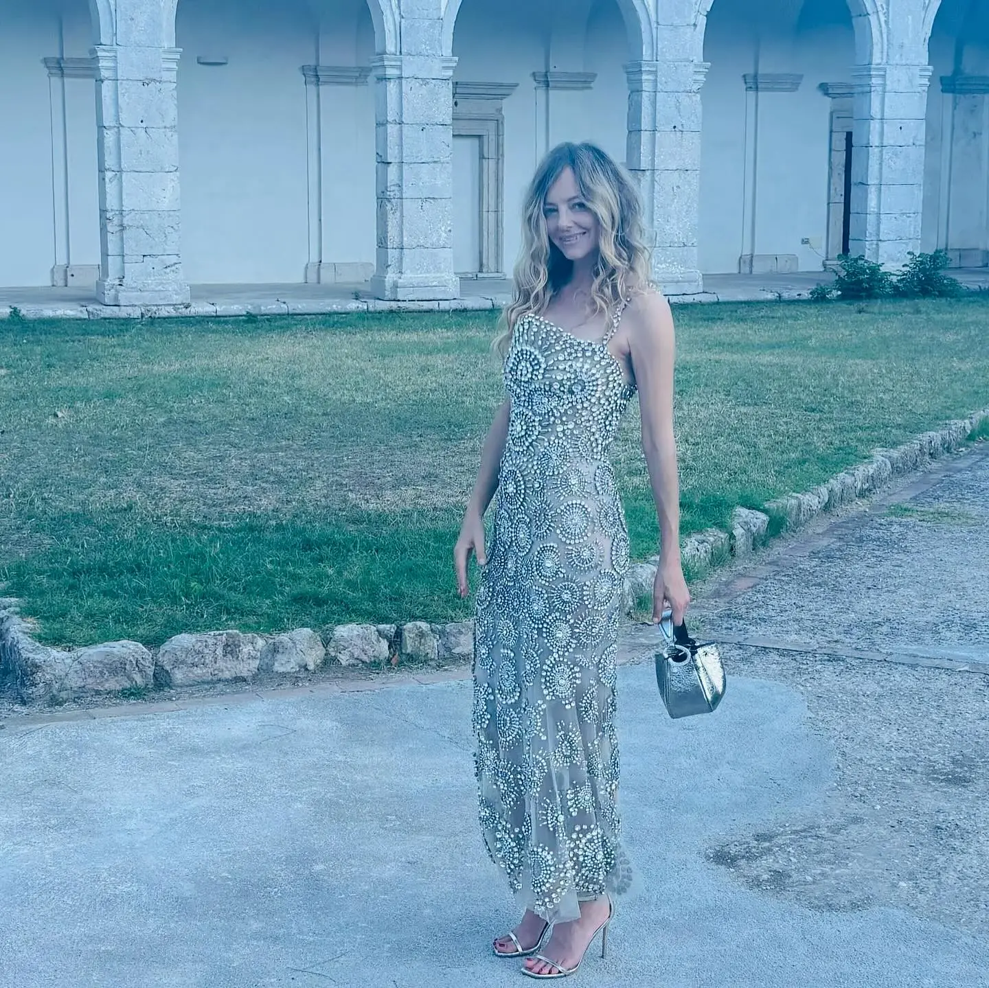 Bijou Phillips smiling in an embellished gown and heels, holding a small silver purse.