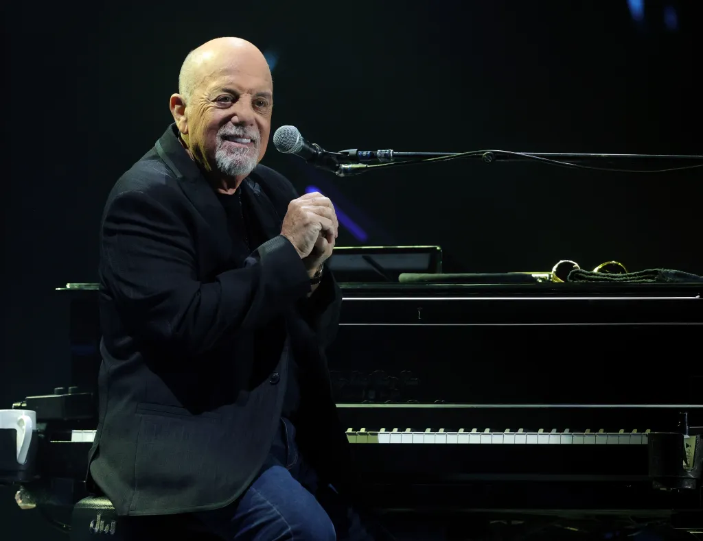 Billy Joel performing at Allegiant Stadium, seated at a piano with his hands clasped and smiling towards the audience.