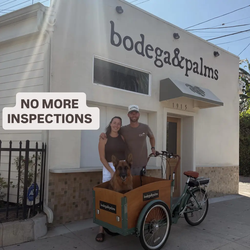 Two people and a German Shepherd in a cargo bike stand in front of "bodega&palms" store.