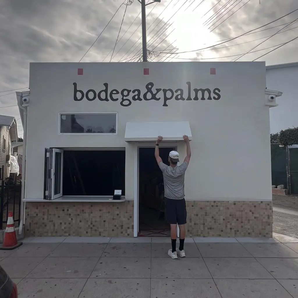 A person adjusting an awning above the entrance of the "bodega & palms" store.