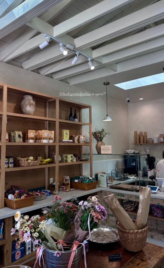 Interior of Bodega and Palms, a market with wooden shelves stocked with various products, fresh flowers on a counter, and a coffee bar in the background.
