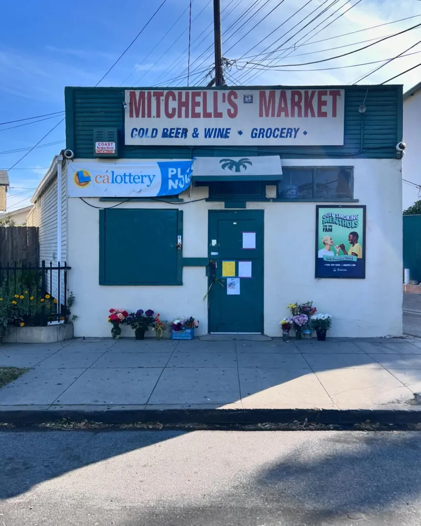 Exterior of the historic Mitchell's Market, now housing Bodega and Palms, with flowers laid on the sidewalk.