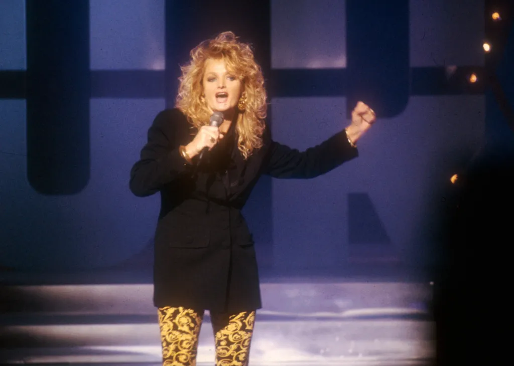 Bonnie Tyler singing into a microphone on stage, wearing a black jacket and patterned yellow and black pants.