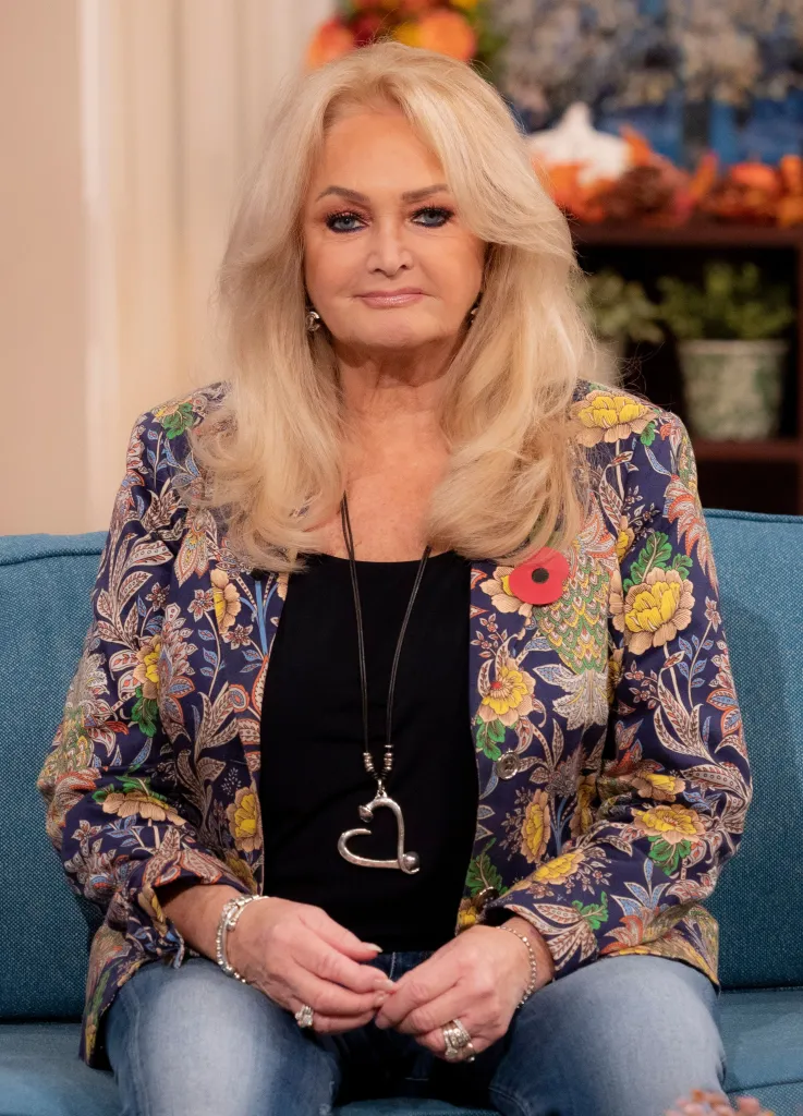 Bonnie Tyler sitting on a blue couch, wearing a black shirt, a floral blazer, and blue jeans, with a poppy pinned to her blazer.