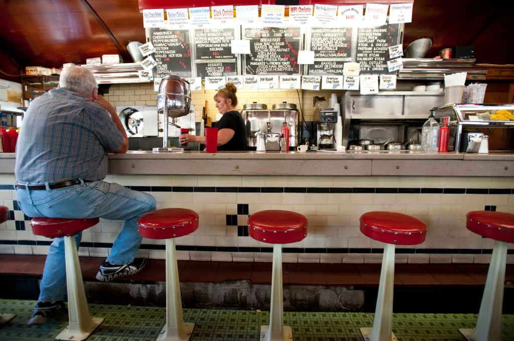 A man sitting at the counter of a diner, facing a woman behind the counter who is filling a cup.