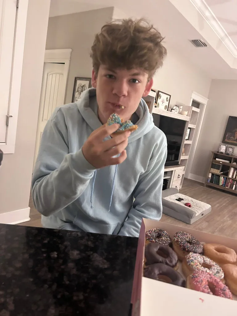 Brandon Baker eating a donut with sprinkles from a box of assorted donuts.