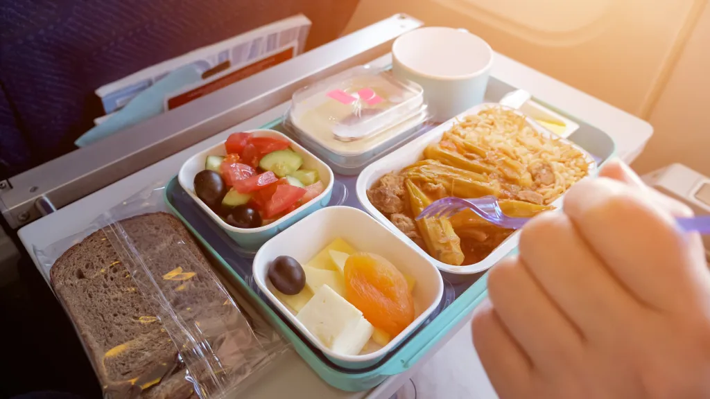 A person eating an airline meal from a tray, which includes bread, salad, rice with meat and vegetables, cheese, and dried apricot.
