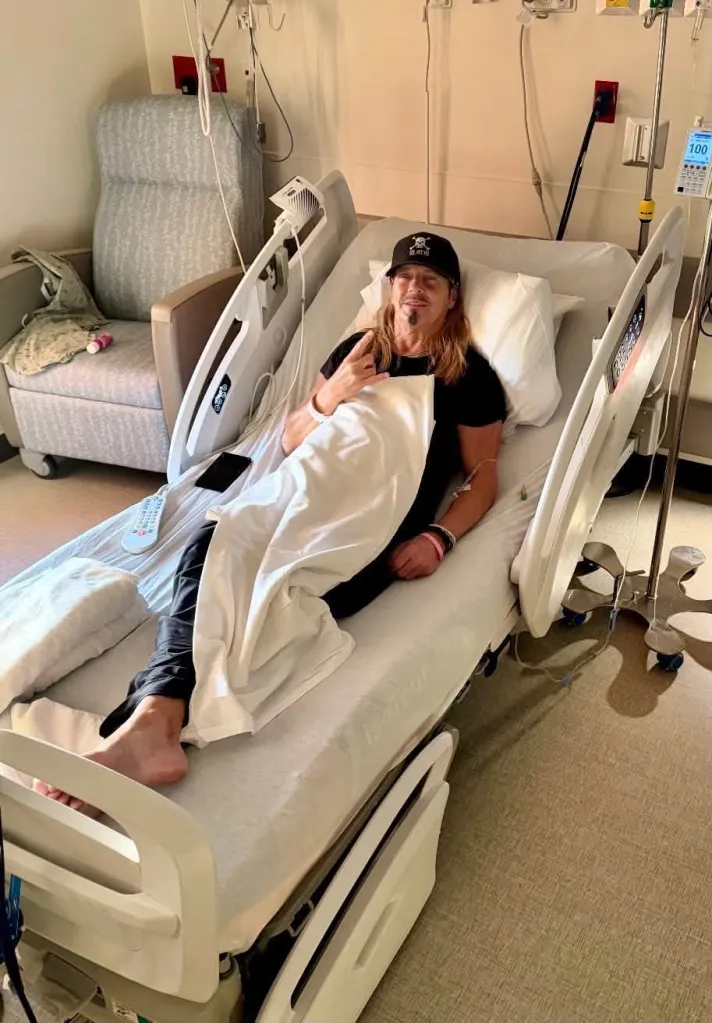 Bret Michaels lying in a hospital bed, making a rock and roll hand gesture.