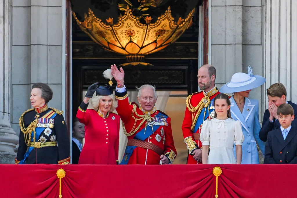 King Charles, in red military jacket, and Queen Camilla, in a red coat and black hat, wave from the Buckingham Palace balcony with Queen Anne in a black military jacket. Prince William in a red military jacket, Princess Charlotte in a blue dress, Kate Middleton in a blue and white coat and blue and white hat, Princes George and Louis in navy blue suits.