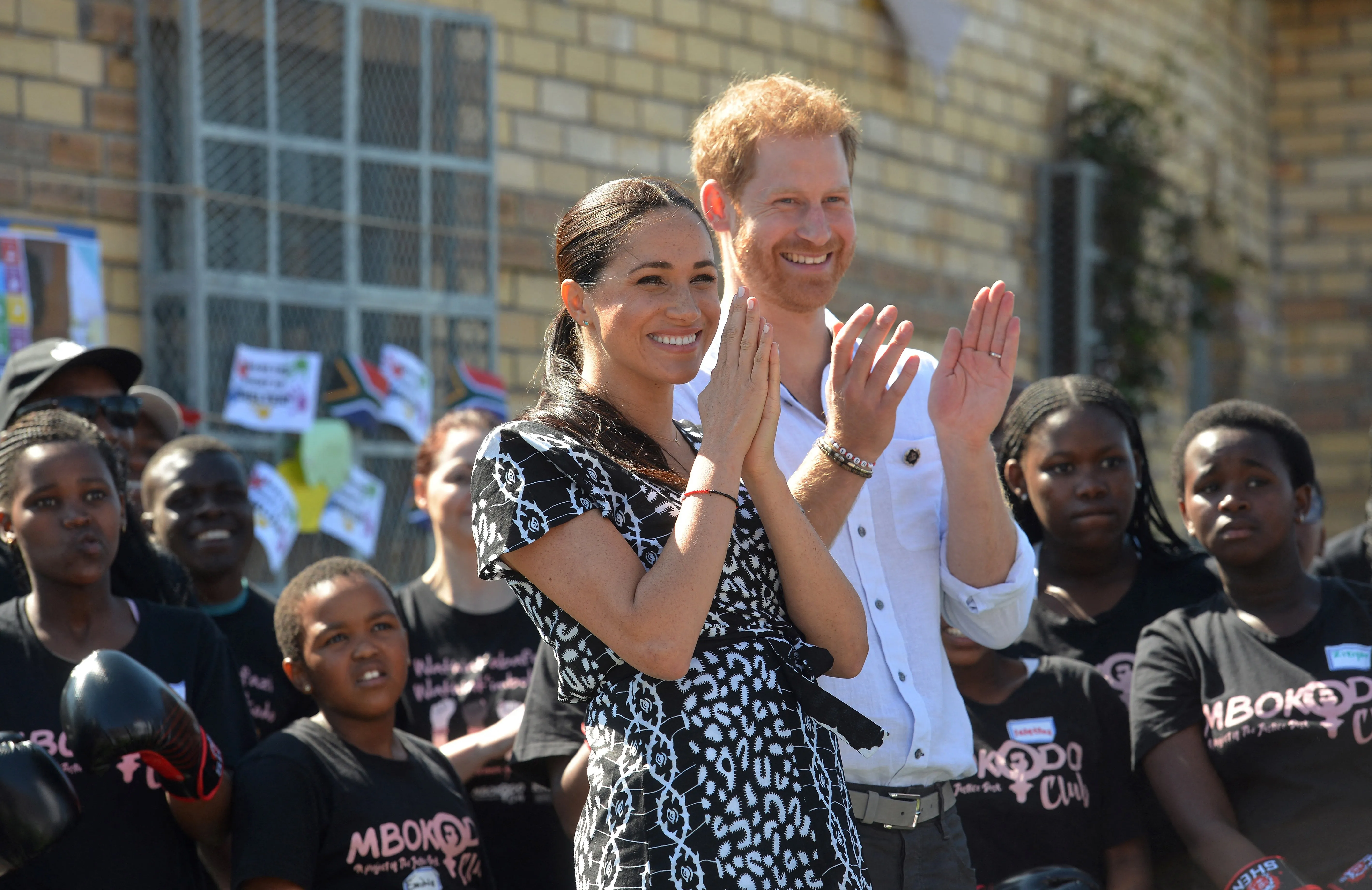 Meghan Markle and Prince Harry clapping with children, some wearing boxing gloves, in the background.