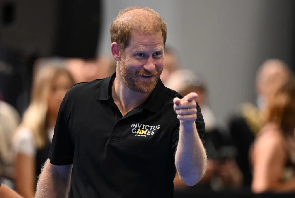 Prince Harry in a black polo shirt with "Invictus Games" logo, smiling and pointing forward.