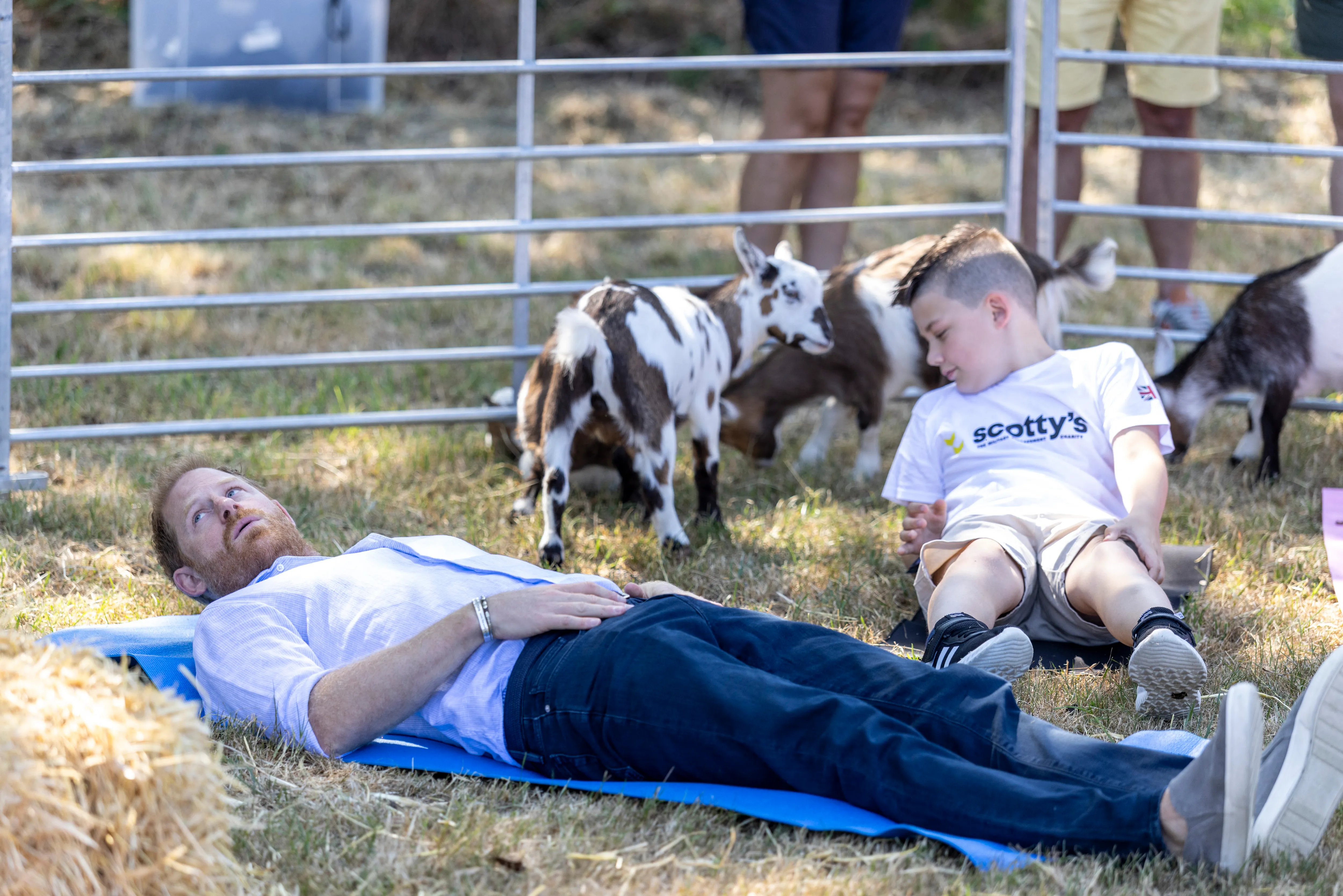 Prince Harry and a child lie on yoga mats on the grass with goats behind them.
