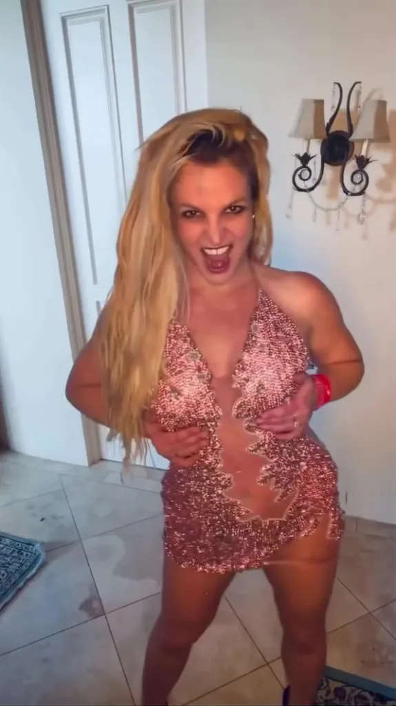 Britney Spears in a sparkly rose gold halter mini-dress with sheer cutouts, with an open-mouth laugh, hands at her chest, in a tiled room with a wall sconce.