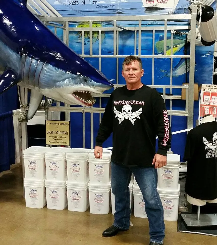 A man in a "Shark Tagger" shirt standing next to a large shark taxidermy mount and a stack of white buckets.
