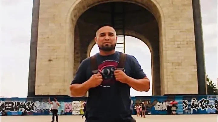 Javier Becerril, a man with a beard and dark t-shirt, wearing a backpack, stands in front of a stone archway with graffiti-covered walls below.