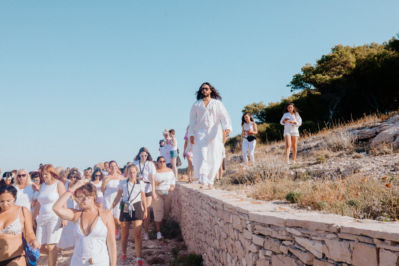 Jared Leto in a long white robe, wearing sunglasses and walking on a stone wall surrounded by followers dressed in white.