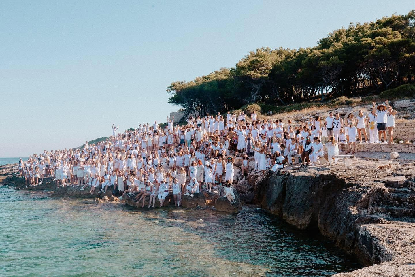 Large group of people dressed in white on a rocky coastline with trees and water.