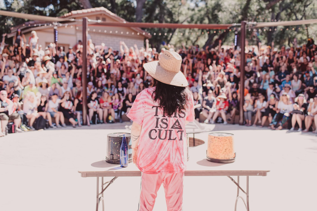 Person in a "THIS IS A CULT" t-shirt speaking to a large crowd.