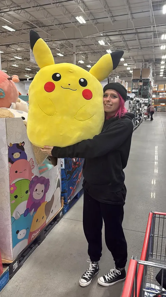 Woman with pink hair hugging a large Pikachu plush toy.