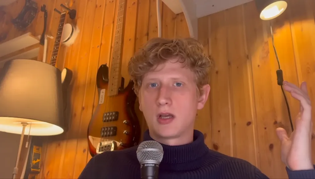 A young man with curly blonde hair and a dark blue turtleneck speaking into a microphone, with a bass guitar, acoustic guitar, and lamps on a wooden wall behind him.