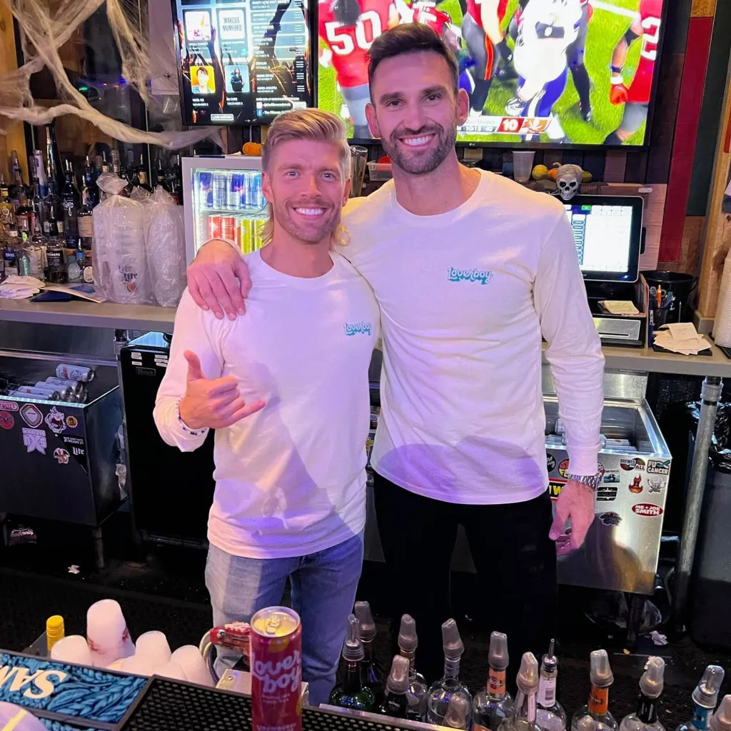 Carl Radke and Kyle Cooke standing behind a bar, both wearing white long-sleeved "Loverboy" shirts.