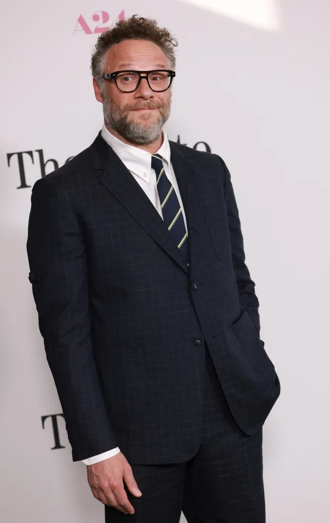 Seth Rogen in a dark checkered suit and striped tie, with a beard and glasses, attending the premiere of "The Invite."