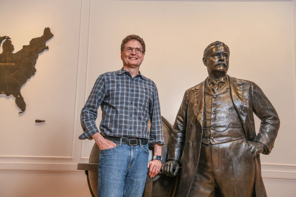 Charlie Melcher standing next to a bronze statue of Teddy Roosevelt.
