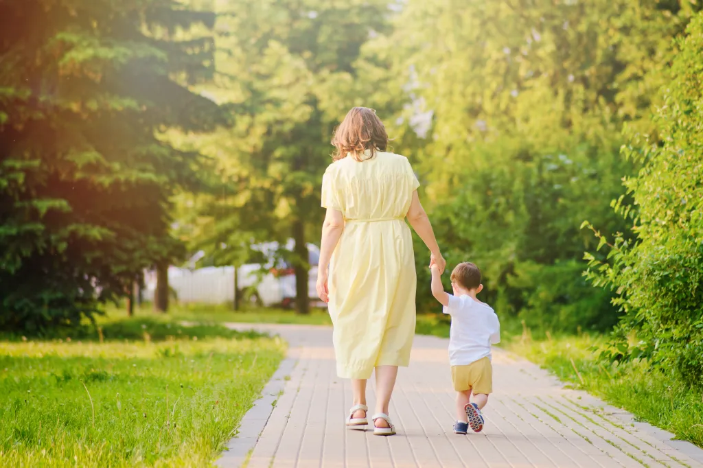 A mother and a two-year-old child holding hands and walking on a path in a park on a sunny day.