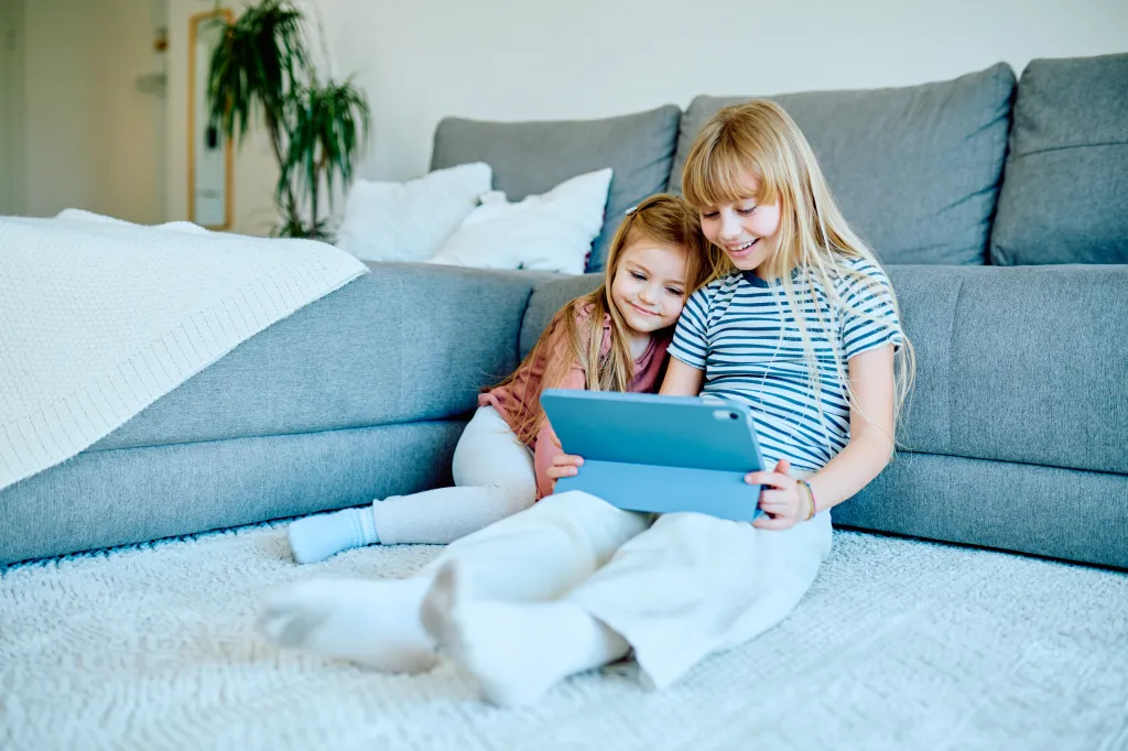 Two young girls, sisters, sitting on the living room floor, smiling and watching a digital tablet together.