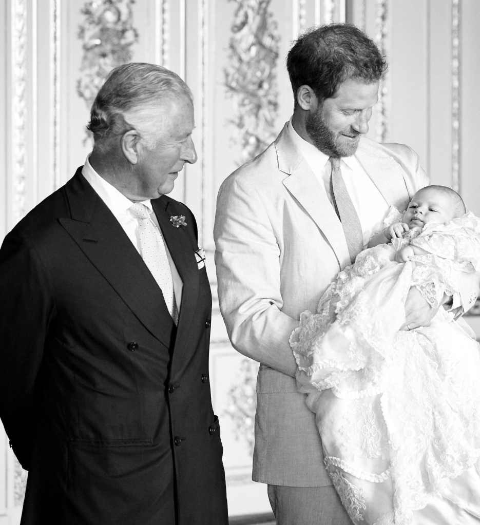 Black and white photo of Prince Harry, in a grey suit, holding baby Archie, in a christening robe, while King Charles III, in a dark sujt, looks on.