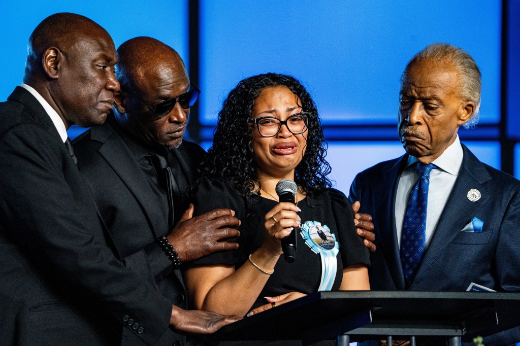 Christine Wonsley speaks into a microphone while being consoled by Ben Crump, Elmore Wonsley, and Rev. Al Sharpton.