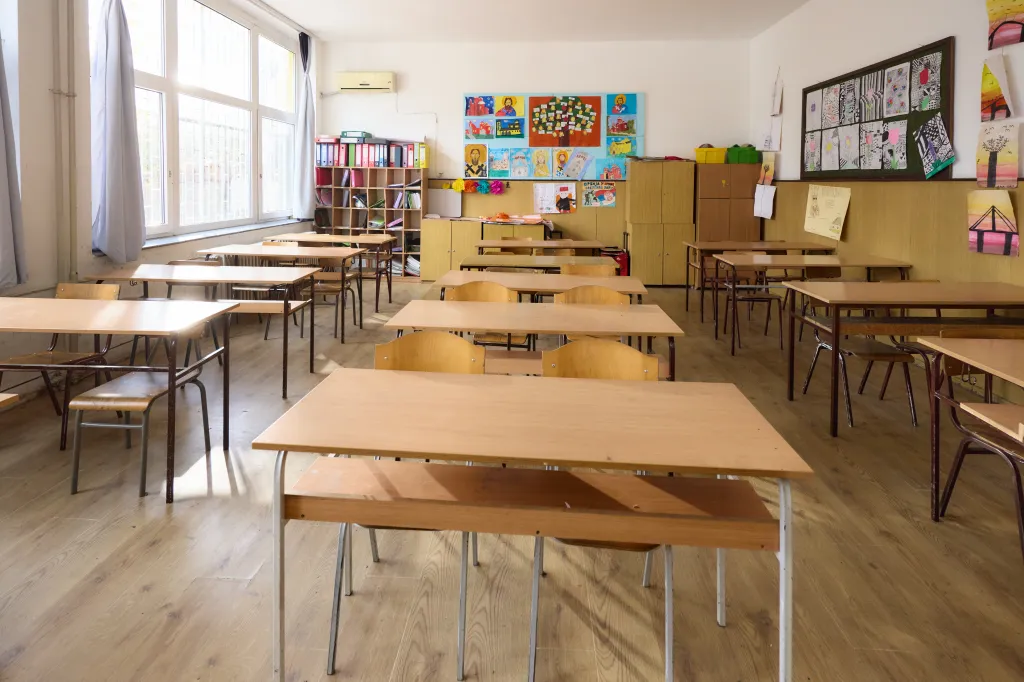 An empty elementary school classroom with desks and chairs facing a wall adorned with children's artwork and shelves of binders.