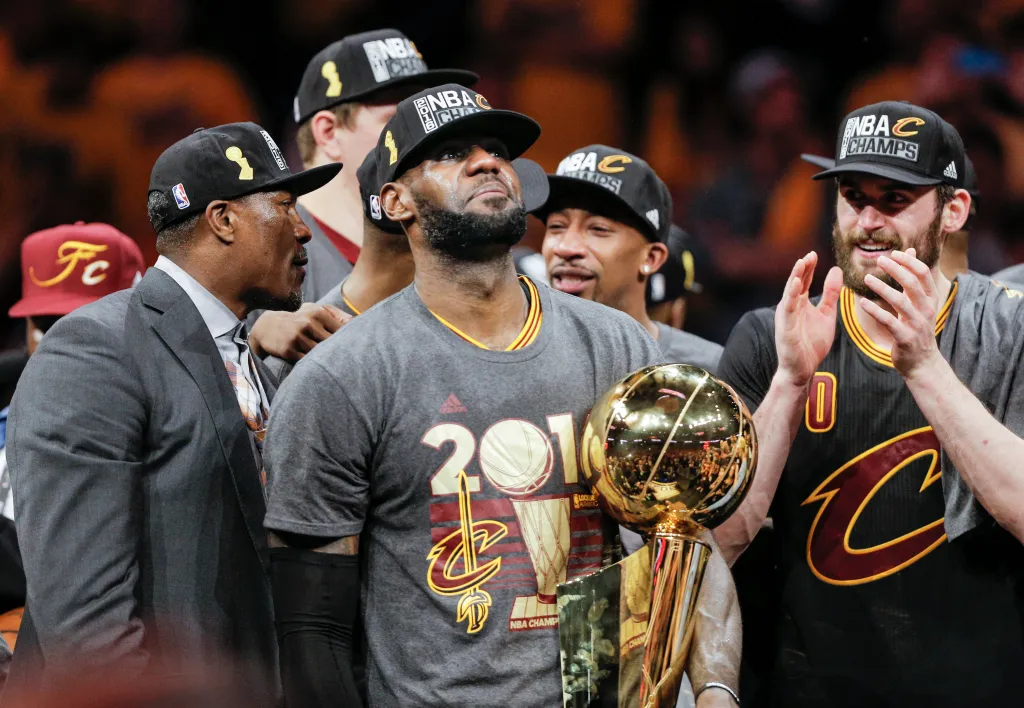 LeBron James holding the Larry O’Brien NBA Championship Trophy while wearing an NBA Champions hat and a "2016 NBA Champs" shirt.
