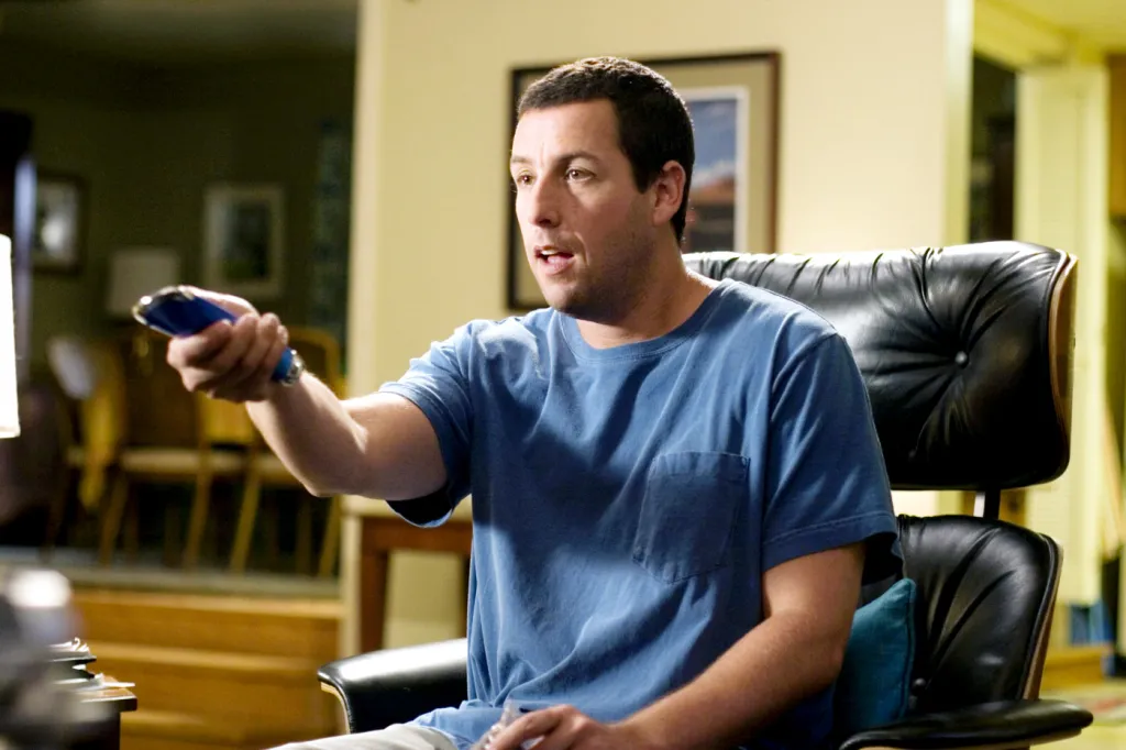 Adam Sandler in character, sitting in an Eames chair and pointing a remote control.