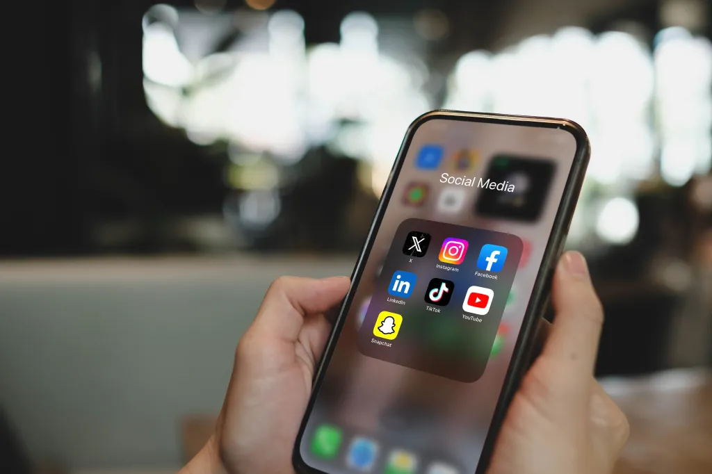 A close-up of an iPhone 15 Pro display showing social media application icons, including X (formerly Twitter), Instagram, Facebook, LinkedIn, TikTok, YouTube, and Snapchat.