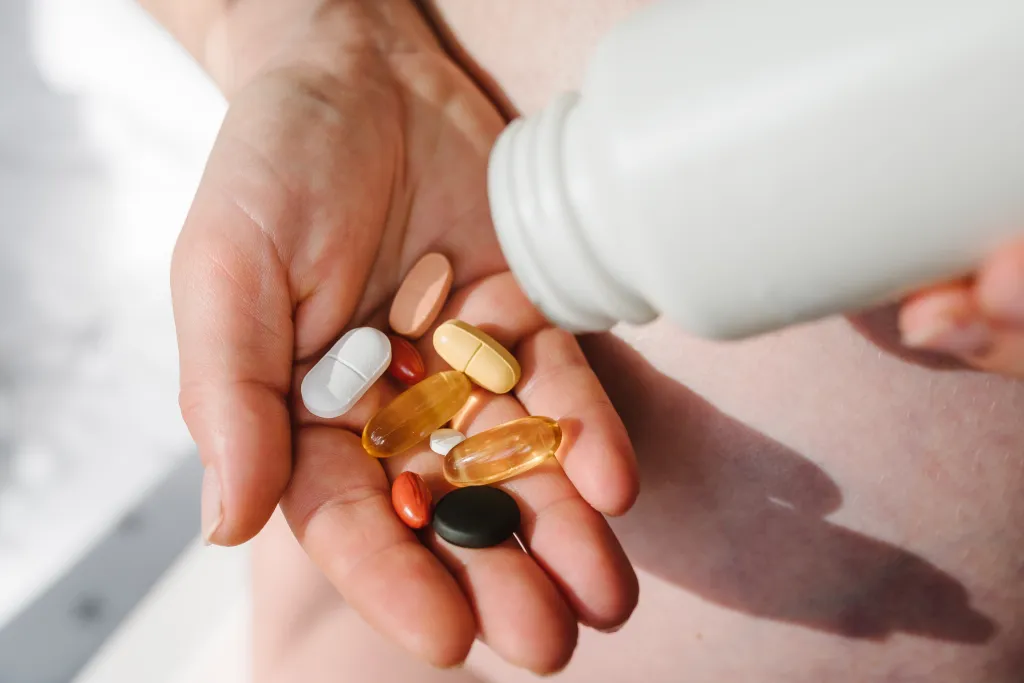 A hand holds a variety of colorful supplements, including omega-3, multivitamins, vitamins B, C, D, collagen tablets, probiotics, and iron capsules, with a white bottle in the background.