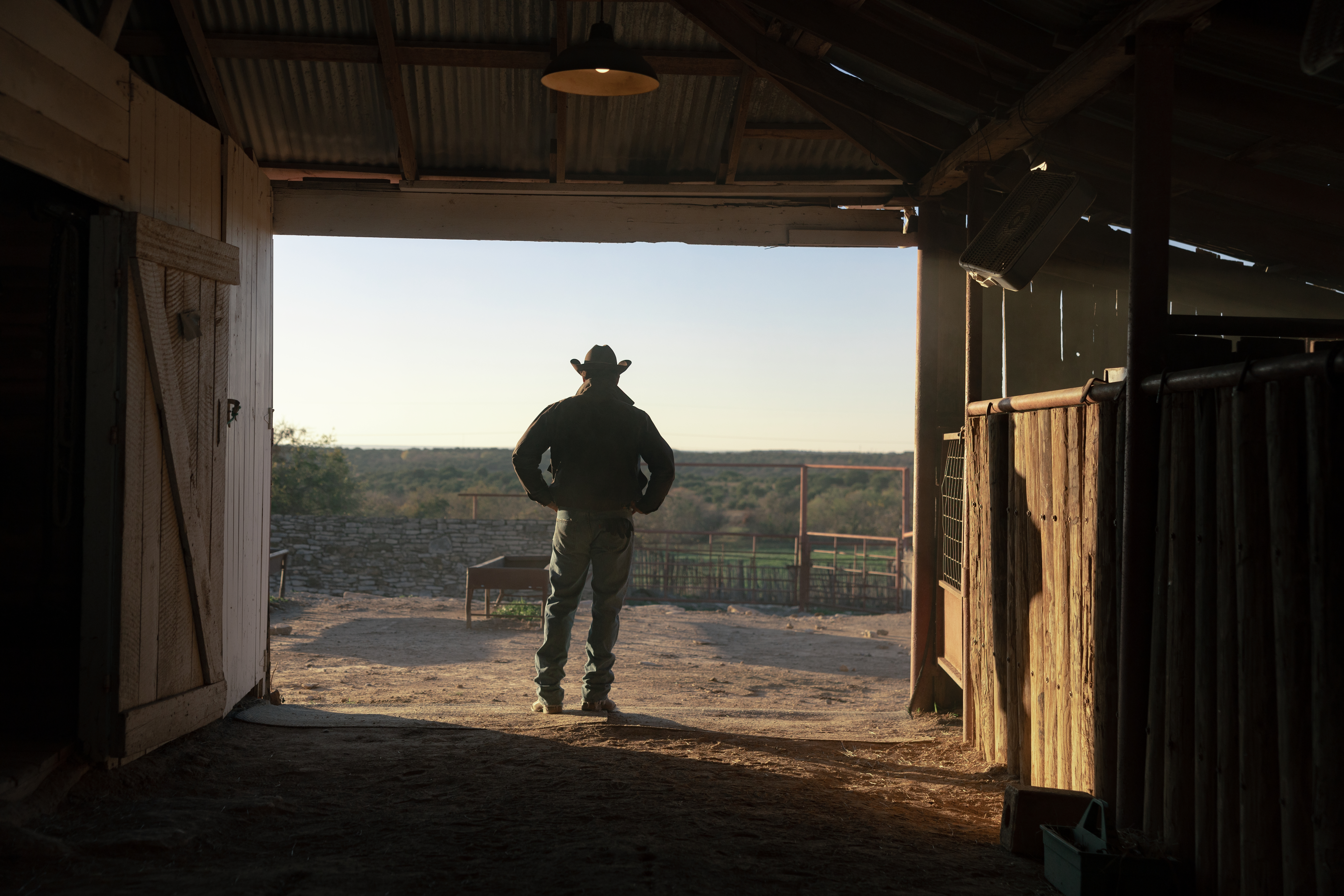 Rip Wheeler in a cowboy hat stands in a barn doorway, looking out at the ranch.
