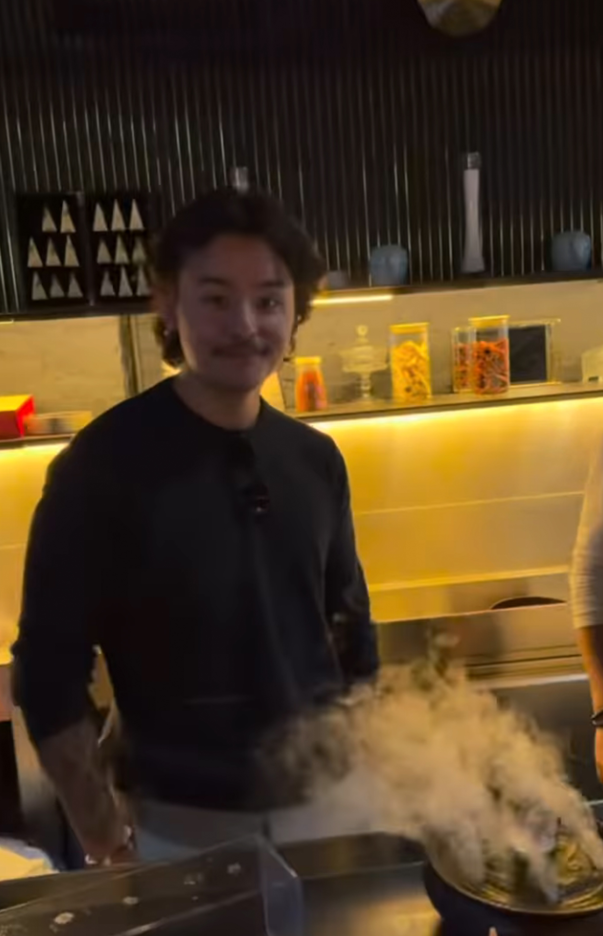 Collin Gosselin smiles, wearing a black shirt, with white smoke coming from a dish in front of him.