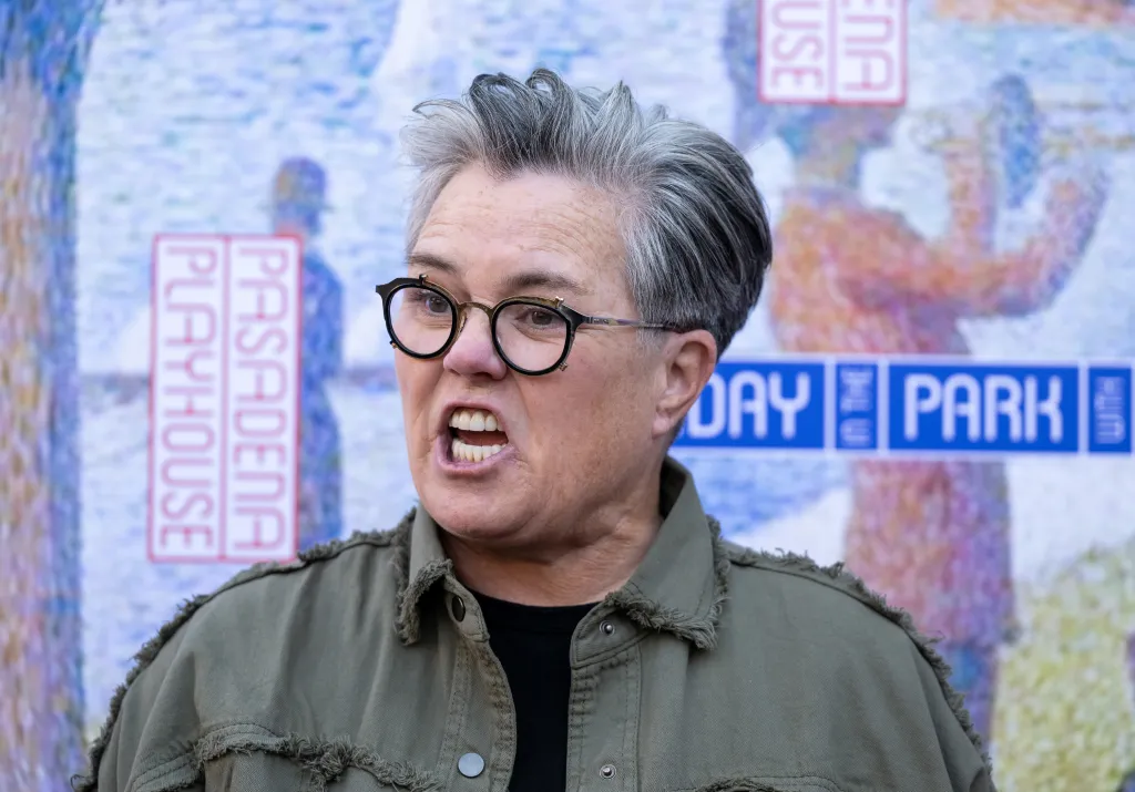 Comedian Rosie O'Donnell with gray hair and glasses, wearing a green jacket, looking forward with her mouth open as if speaking.